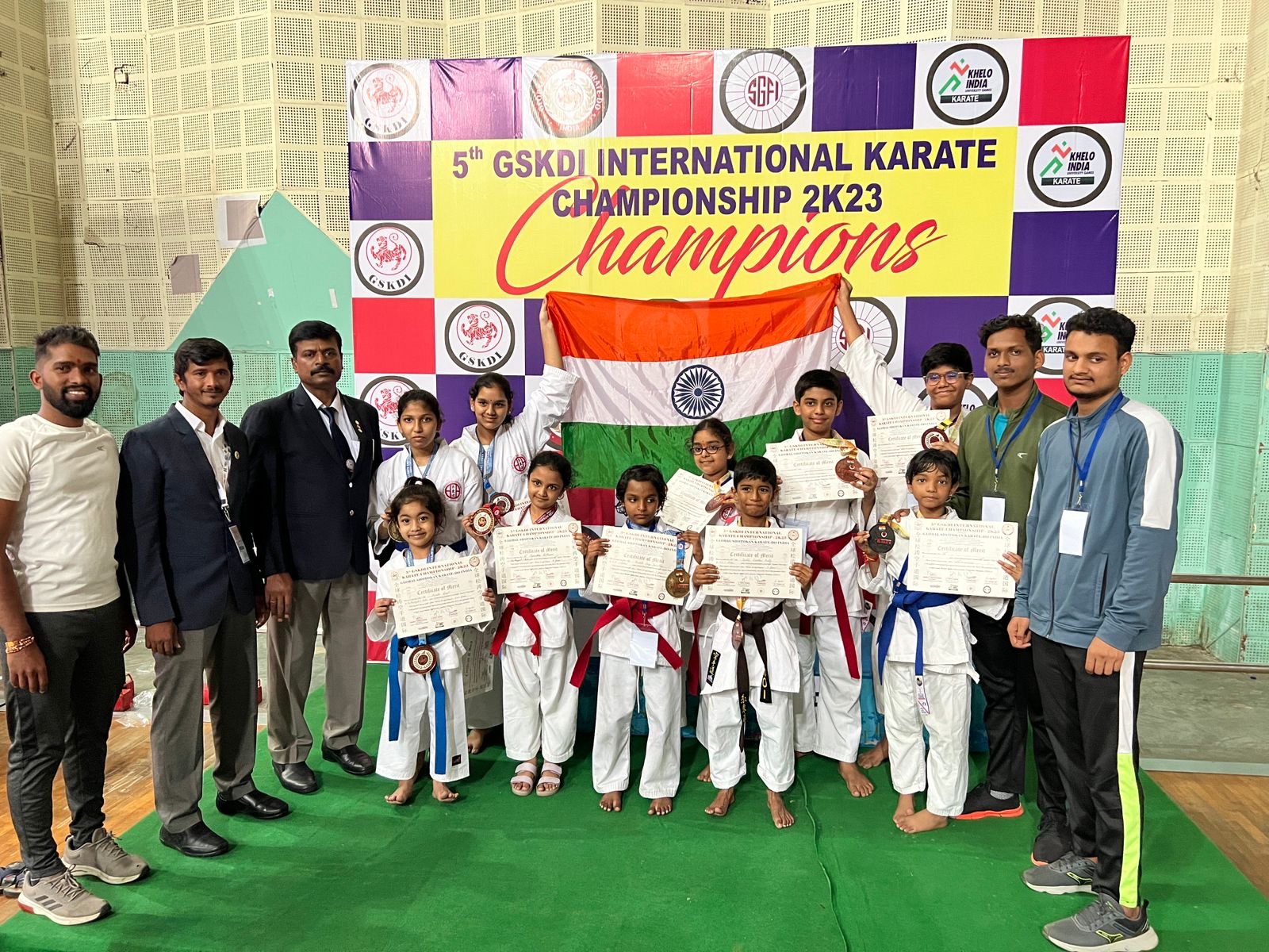 United's young karateka in white gis holding medals, certificates and the Indian flag under the 5th GSKDI International Karate Championship 2K23 Champions banner, with their coaches.