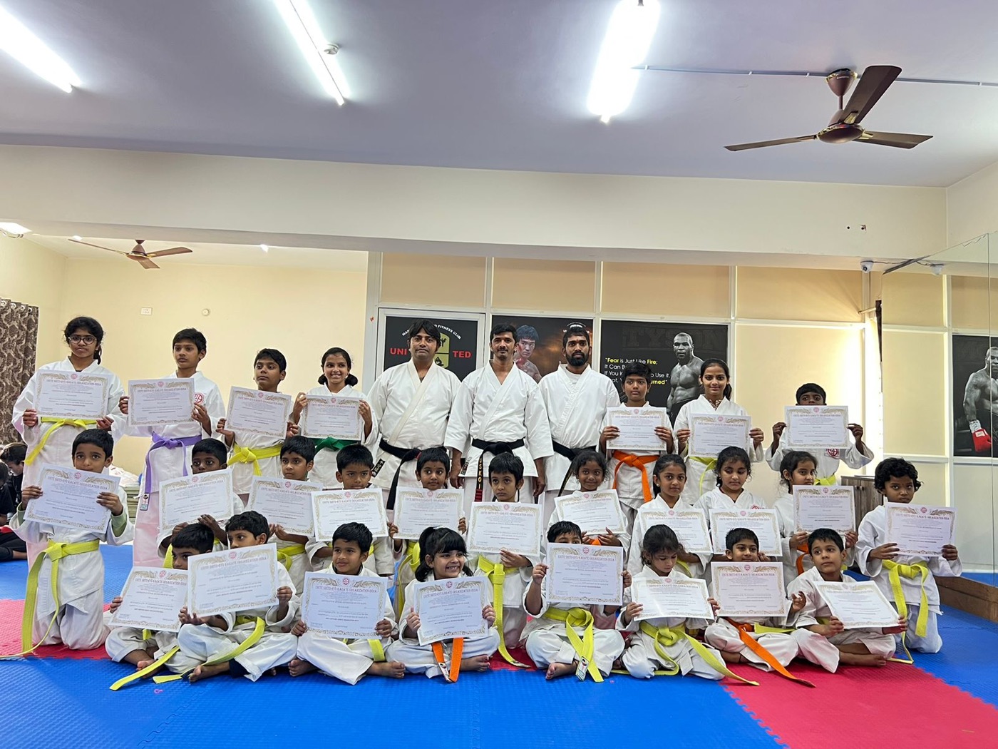 Kids' batch on the blue and red mats holding grading certificates, with black-belt instructors standing behind them.